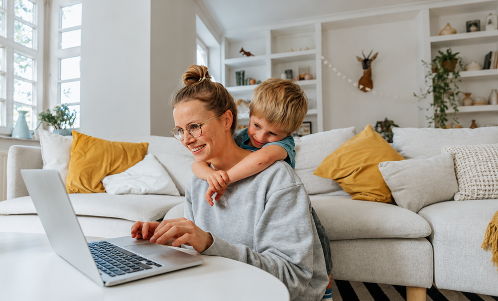 Woman using a laptop at home while a young child hugs her from behind.